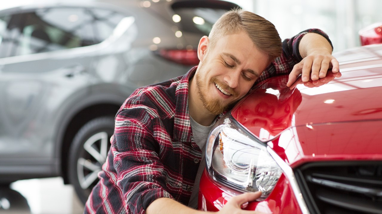 Man in flannel shirt hugging the font end of a new red car in a showroom - car insurance in louisiana - kelly lee insurance